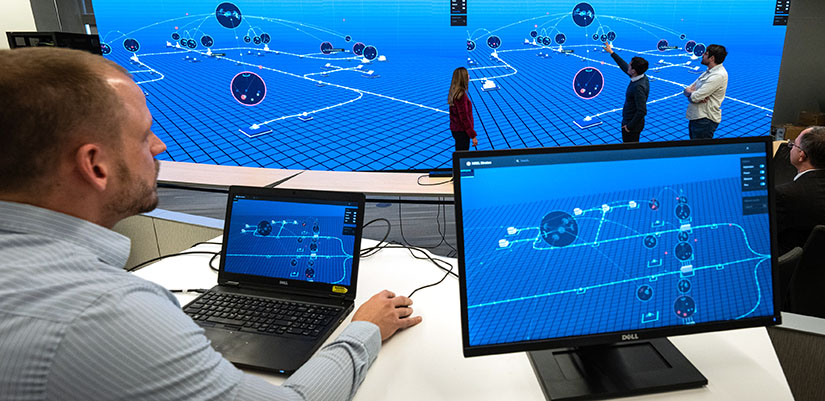 Person navigates laptop while screen displays on a large screen where other stand in front and point.