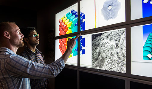 two researchers facing a wall of computer monitors in a lab