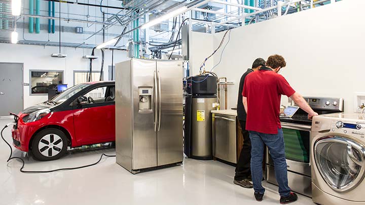 Two men looking down at a laptop while standing near kitchen appliances and a charging electric vehicle.