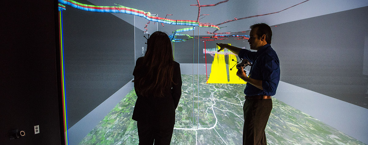 A man and woman standing in front of a large, color 3D visualization screen that spans the height and width of the wall they are facing.