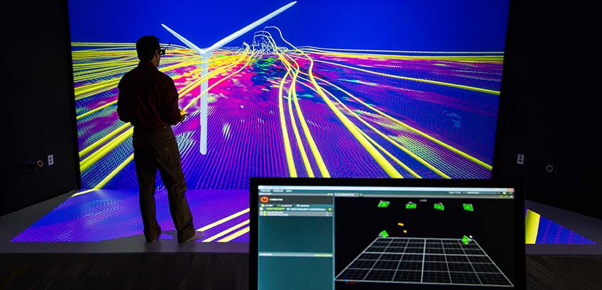 Photo of a researcher viewing a 3-D model of a wind turbine simulation with a computer monitor in the foreground displaying a program.