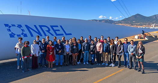 Group of people stand outside NREL campus in front of wind turbine blade.