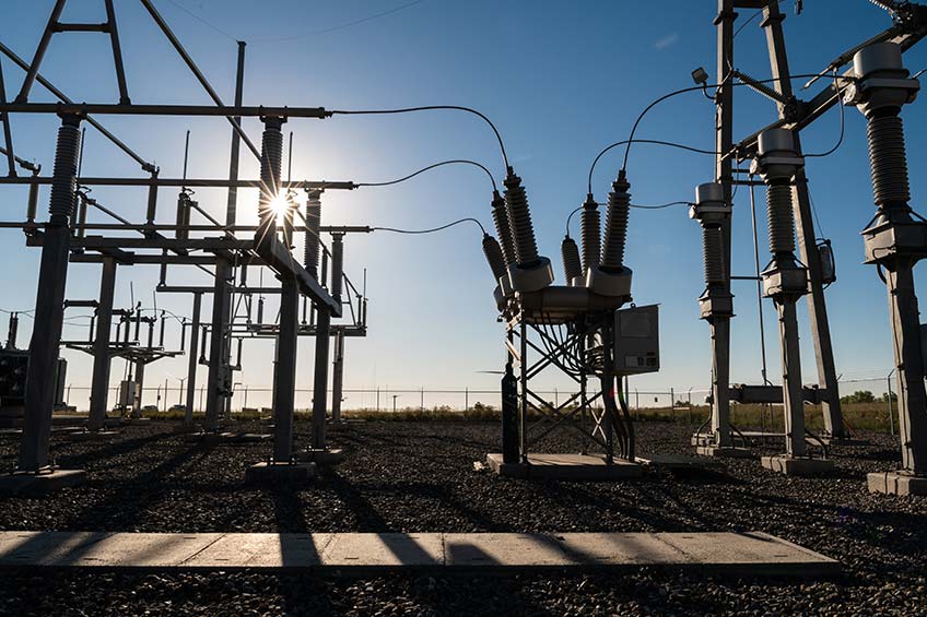 The sun peaks through a power grid system, casting shadows on the ground.