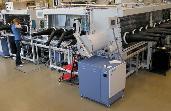 Photo of lab equipment featuring a glass enclosure with many long, black arm gloves, and a female researcher sticking her arm into one of gloves to work on something in the enclosure. 