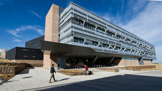 Photo of a three-story, beige, grey, and black building with two people walking in front of it.  