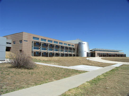 Photo of a three-story, brown building with a silver, cylinder-shaped entrance. 