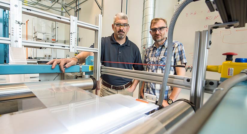 Two scientists stand with roll-to-roll equipment in lab.