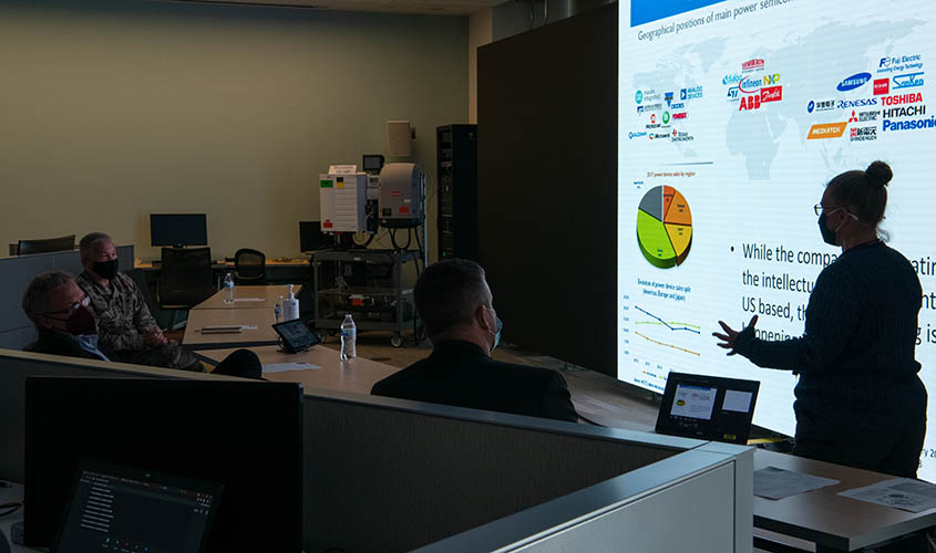 A woman presents in front of a screen to three people in medical masks.