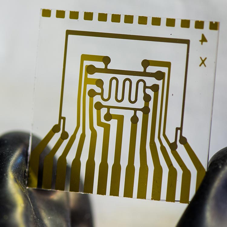 A photo of a glass square held between two fingers. The glass has a pattern of metal contacts that make up a special electrical circuit. This device represents an electrical calcium test glass card used to measure vapor-barrier permeation rates.