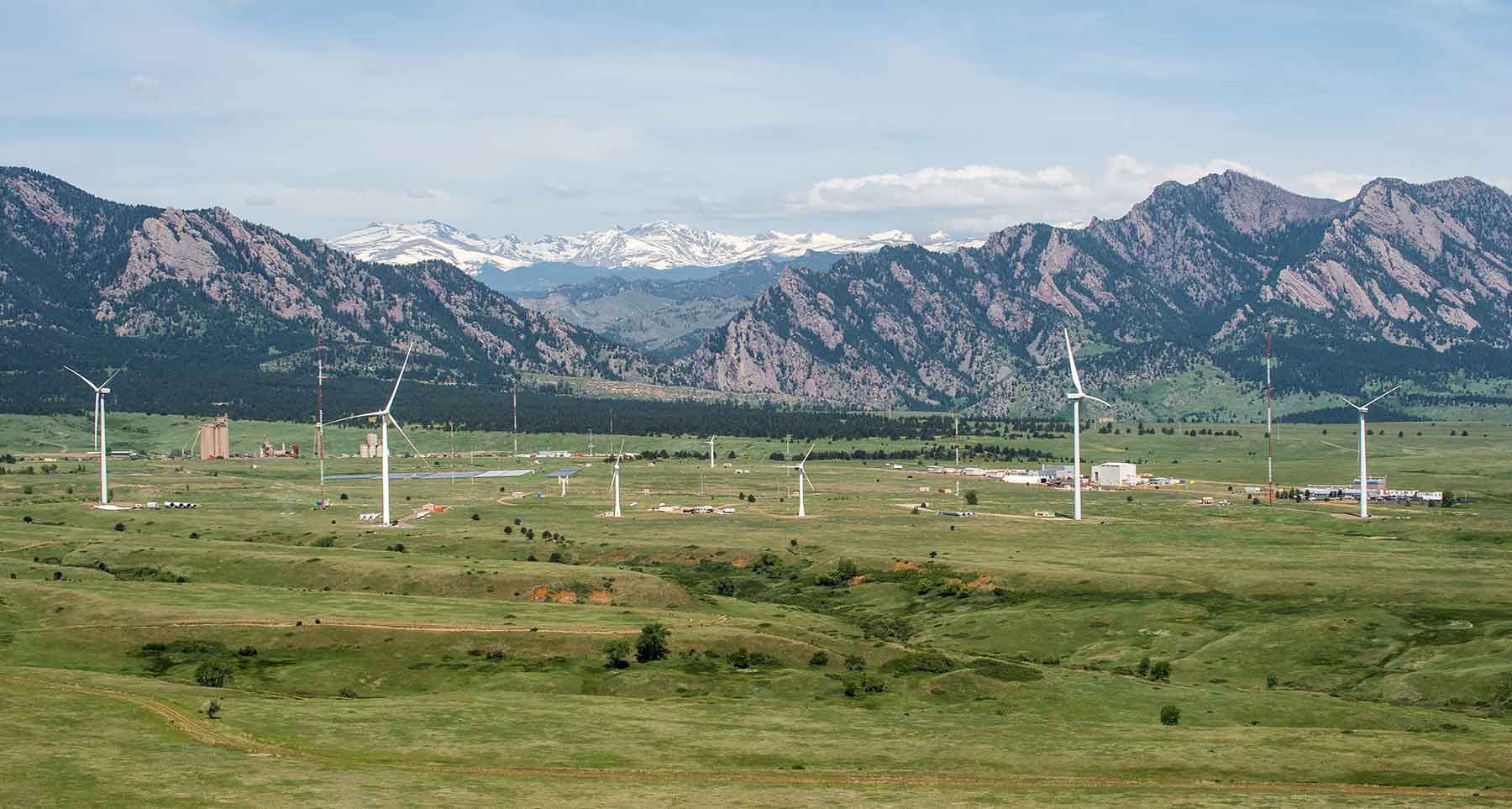 A wide aerial view of a wind turbine testing site and the landscape surrounding it. One large wind turbine is in the foreground with four other wind turbines behind it, as well as several buildings and met towers. Mountains are in the background