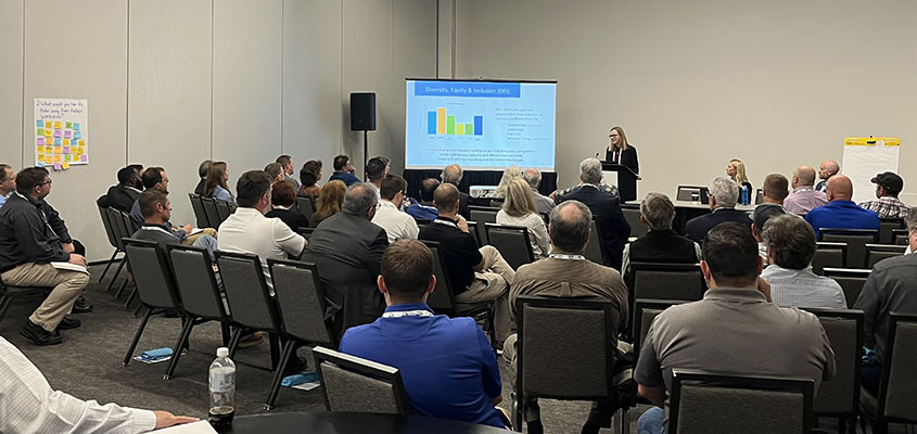 A woman speaking at the front of a conference room full of attendees. Next to her is a large screen showing a graph.  