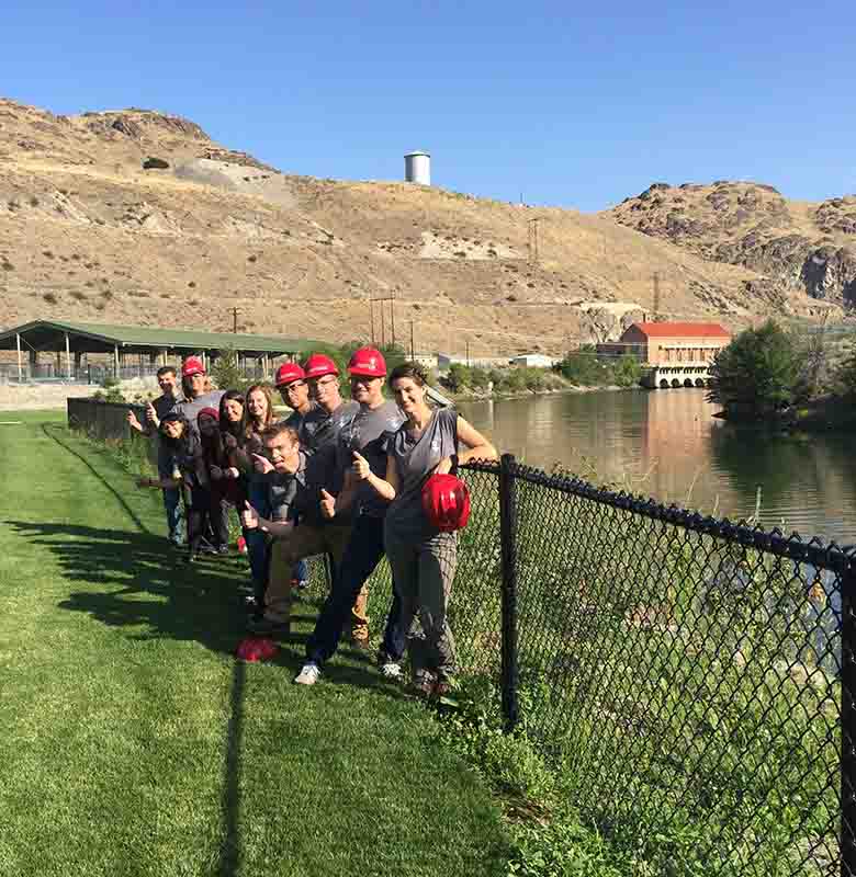 A group of students in hard hats standing in grass and leaning on a fence next to a waterway.