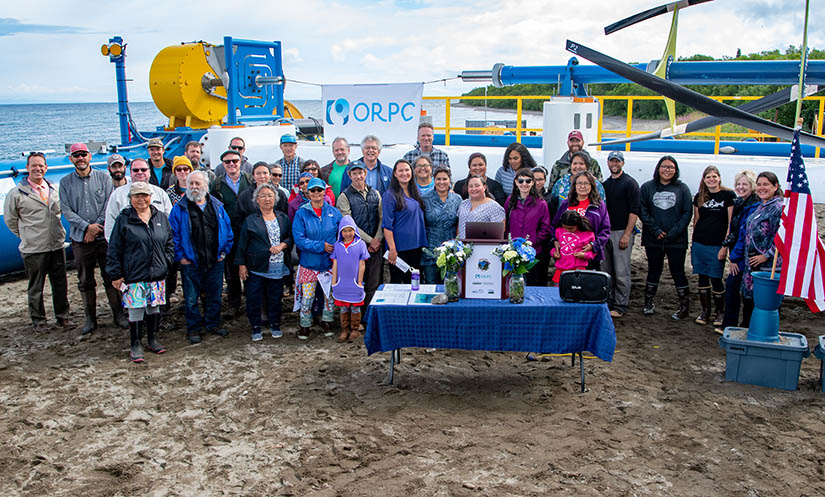 A crowd of people standing on a beach in front of a mechanical device and a river.