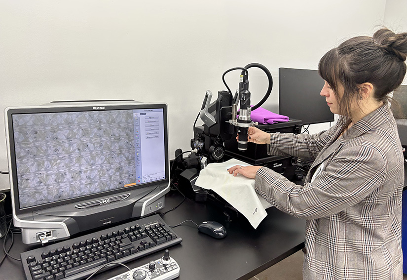 A person examines a fabric under a microscope next to a computer screen.