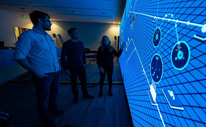 Three people stand in a dark room looking at a large wall display computer screen.