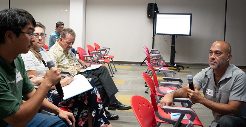 A person who is sitting in a row of chairs speaks into a microphone, while another person kneeling in the row in front of him listens.