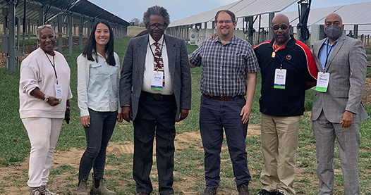 Group of people stand outside in front of solar panel array on ground.