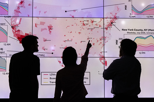 Three silhouetted people looking at a data visualization with lines, charts, and a U.S. map on screens behind them