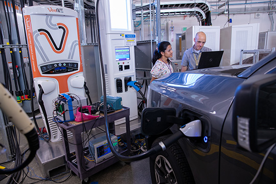 Two people look at a laptop placed on the hood of an EV plugged into a charger in an indoor NREL lab.