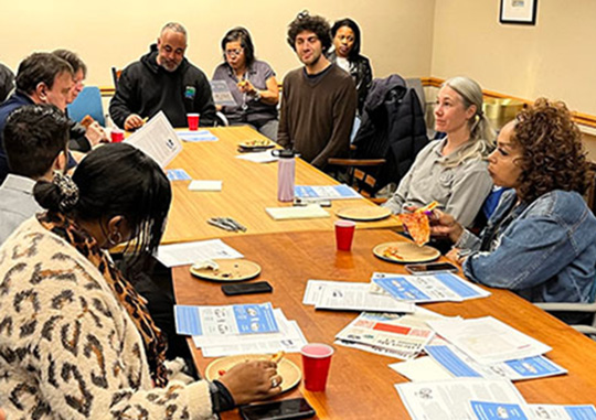 A group of 13 people sitting around a table with flyers and plates of food on it. Two other people are standing, one of whom is speaking.