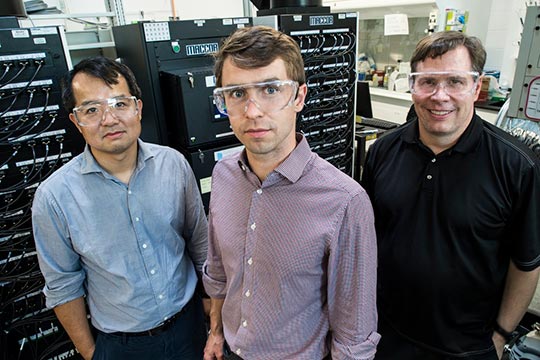 Three men pose for the camera with electronic devices in the background