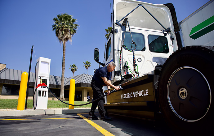 Photo of man standing near an electric vehicle charging station and inserting a charging nozzle into the side of a heavy-duty electric truck.