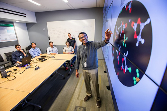 A researcher stands and presents in front of a large screen. Five other people sit and watch the presentation.