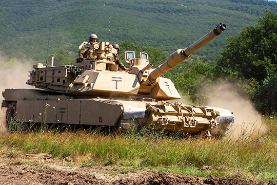 Several U.S. soldiers driving an M1 Abrams tank across a green field, kicking up a cloud of dust.