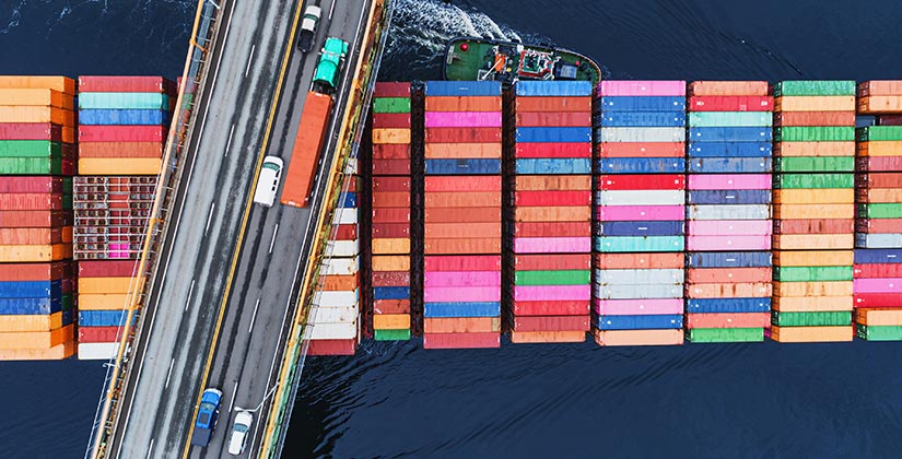 A photo of cars and trucks driving across a bridge as a freight ship with colorful cargo passes underneath.