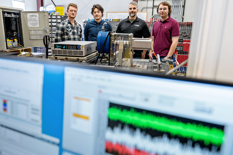 Data on computer screens with four people in the background working on a wave energy converter device.